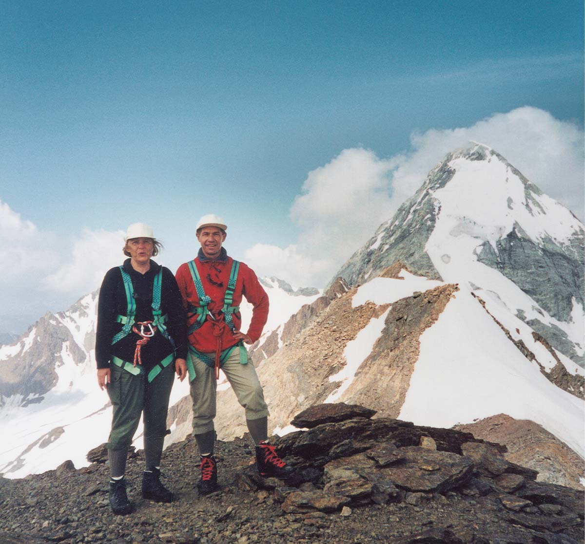 Angela Merkel und Joachim Sauer in Wanderkleidung auf einem Berg, mit Sicherheitsgurten und Helmen, im Hintergrund schneebedecktes Gebirge