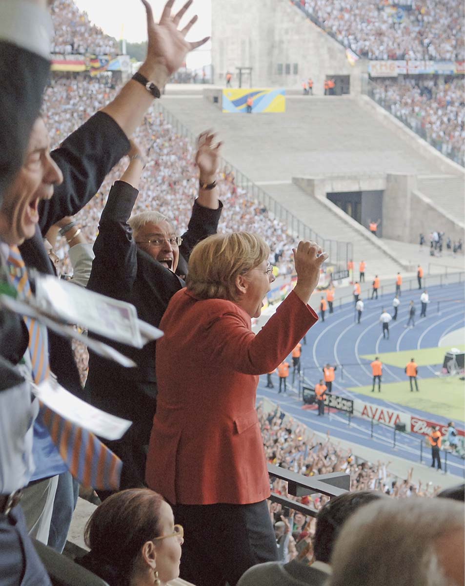 Angela Merkel in einem vollbesetzten Fußballstadion, auf das Spielfeld blickend, die Arme jubelnd hochreißend