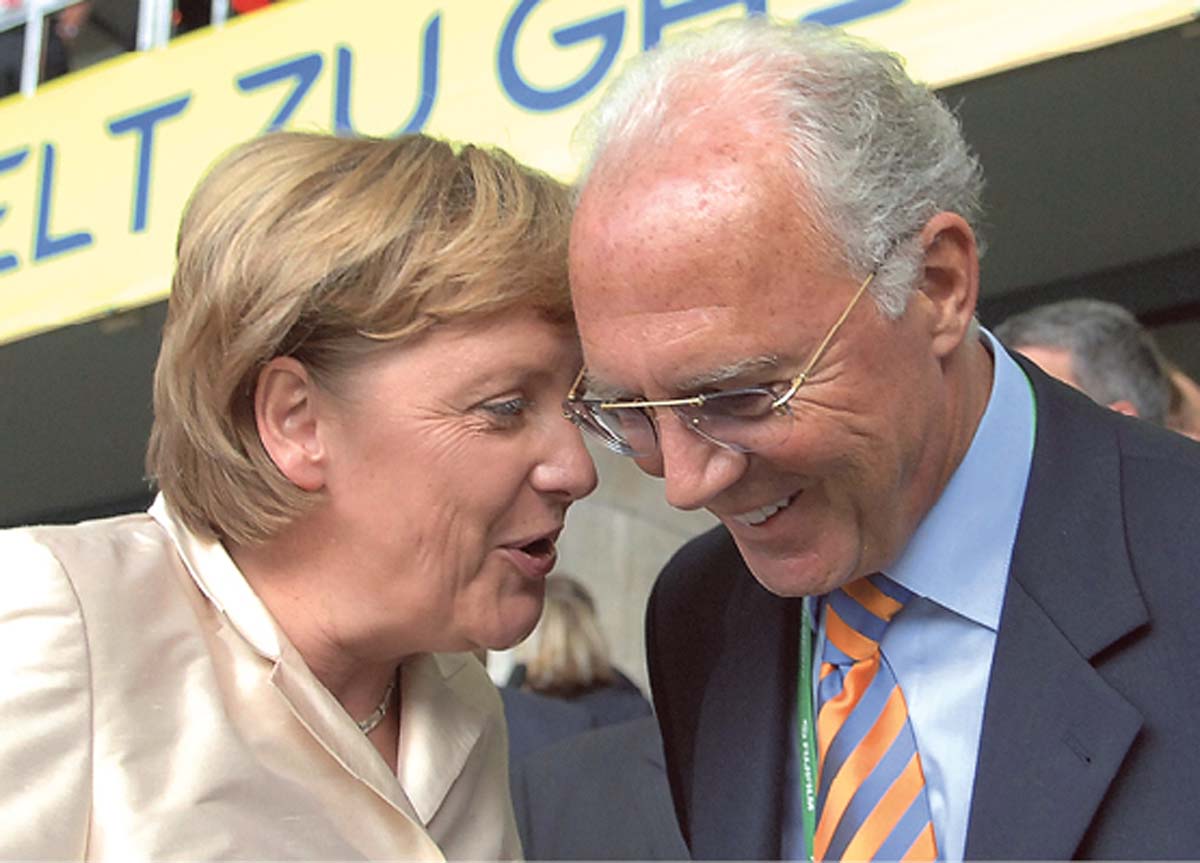 Angela Merkel spricht Franz zu Beckenbauer, der lächelnd zuhört, im Hintergrund erkennbar weitere Fans.