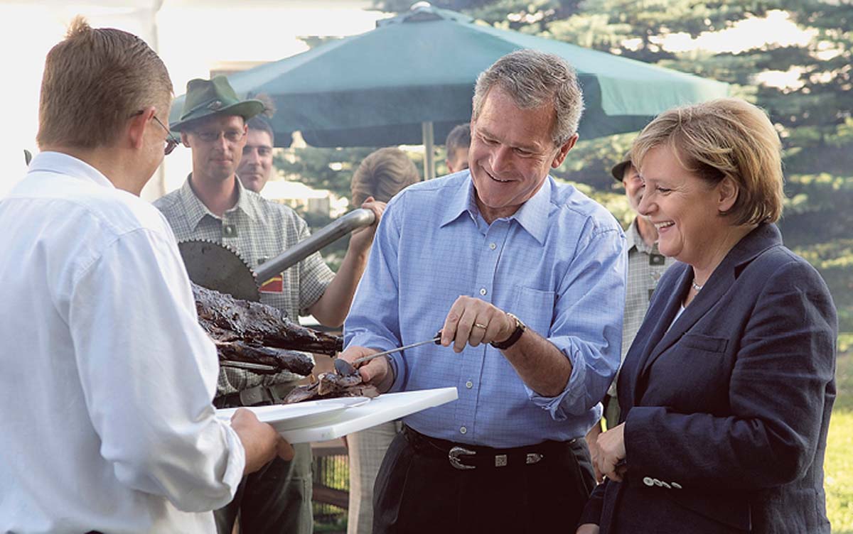 Angela Merkel steht neben Geoge W. Bush, der offenbar mit einem Messer in der Hand in Grillgut sticht, das ihm ein Mann in weißem Hemd hinhält. 