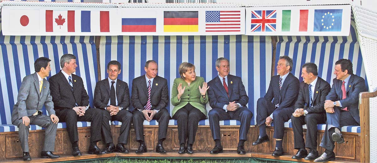 Angela Merkel in der Mitte eines riesigen Strandkorbs sitzend, links und rechts neben sich jeweils vier Männer in Anzügen