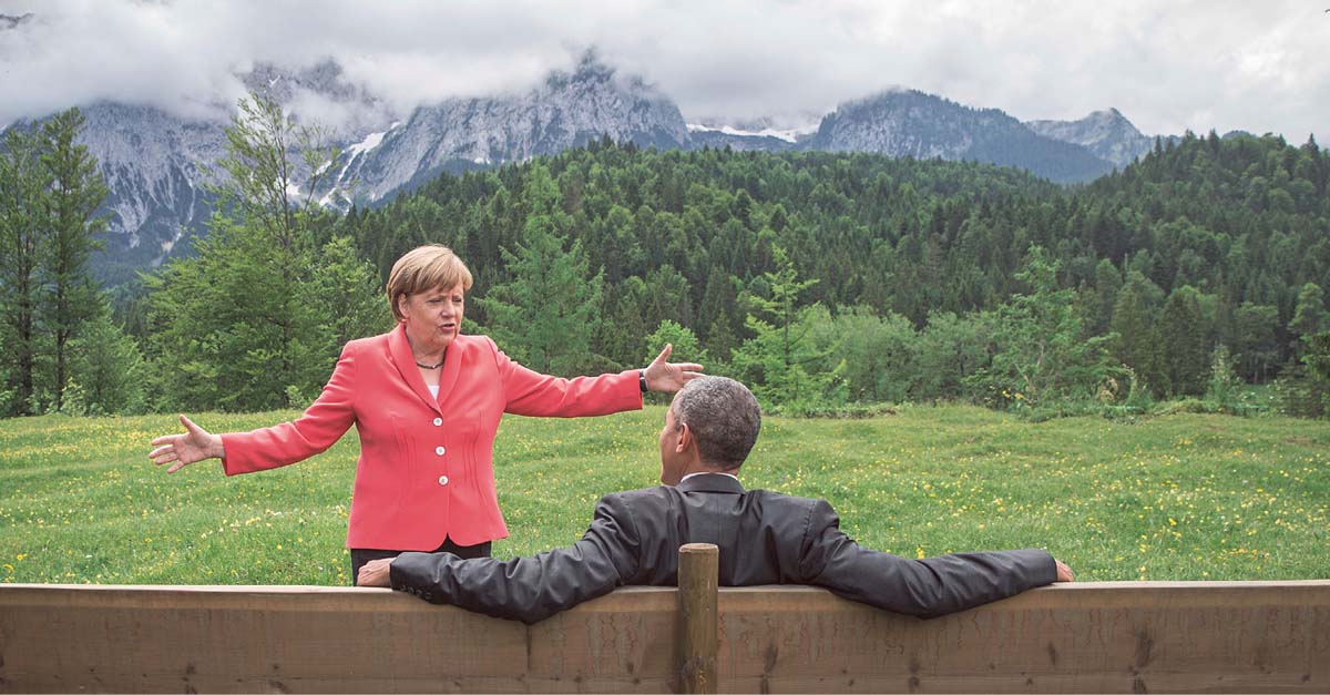 Angela Merkel steht, die Arme ausbreitend, vor dem auf einer großen Holzbank sitzendem Barack Obama, im Hintergrund die Alpen