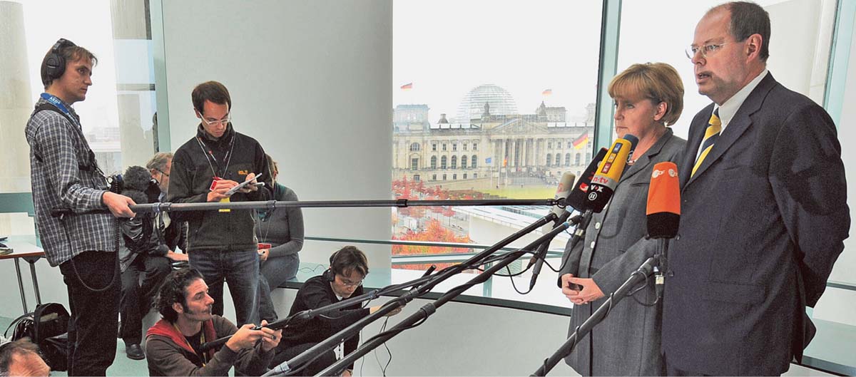 Angela Merkel und Peer Steinbrück stehen vor einer Gruppe Journalisten, die ihnen an langen Stangen befestigte Mikrofone hinhalten. Im Hintergrund ein großes Fenster, durch das man den Reichstag sieht