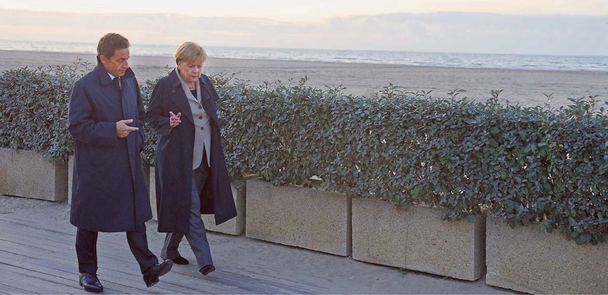 Angela Merkel und Nicholas Sarkozy, beide in dunklen Mänteln, laufen über eine Strandpromenade, vorbei an einer halbhohen Hecke, dahinter der Strand und das Meer.