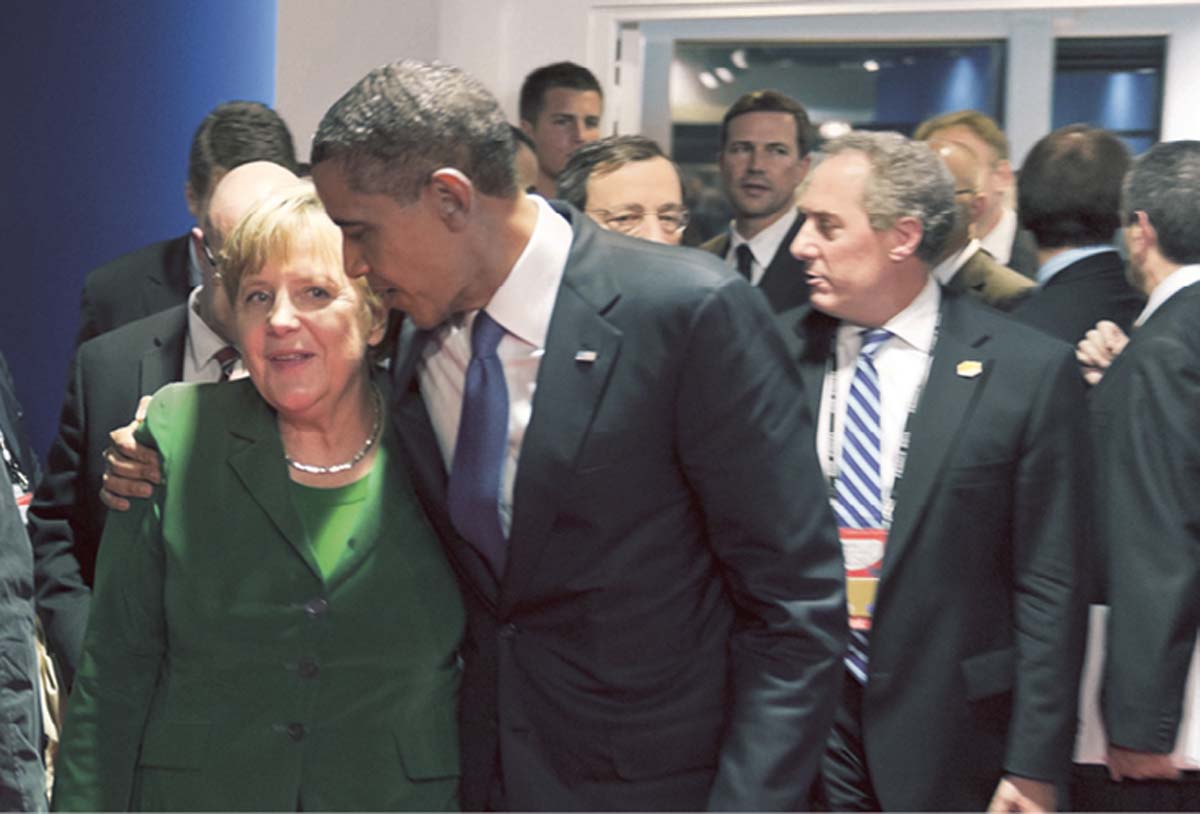 Angela Merkel und Barack Obama laufen vor einer Gruppe Menschen her, er dabei hat seinen Arm um ihre Schultern gelegt und schaut sie an.