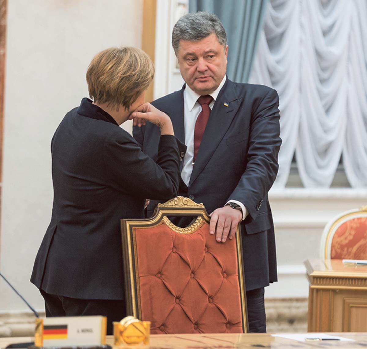 Angela Merkel, von hinten fotografiert, an einem Stuhl lehnend und Peoroschenko anblickend, der zur Seite schaut