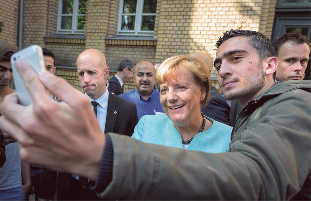 Ein arabisch gelesener junger Mann steht neben Angela Merkel und hält ein Handy vor sich, um ein Selfie zu machen; beide stehen vor einem Gebäude im Freien, hinter ihnen weitere Menschen