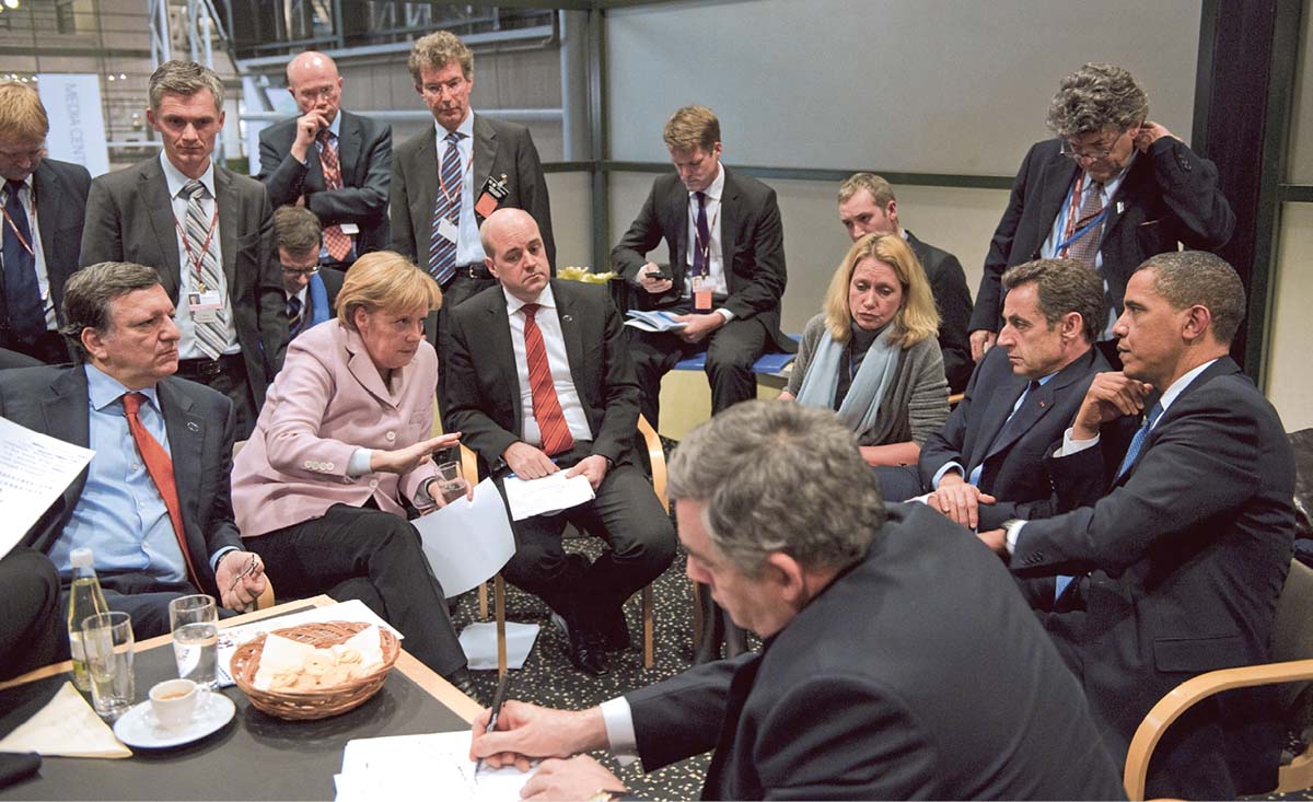 Angela Merkel inmitten einer Gruppe von Männern in dunklen Anzügen, die um sie herum sitzen oder stehen. Außer ihr ist nur noch eine weitere Frau auf dem Foto zu sehen.