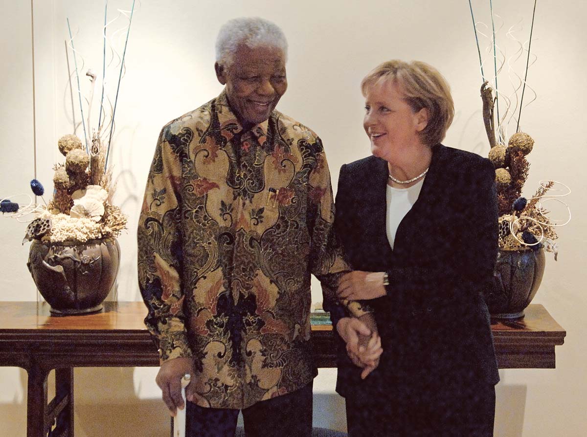 Angela Merkel schaut lächeln den neben ihr stehenden Nelson Mandela an. Im Hintergrund Blumengestecke auf einem Holztisch