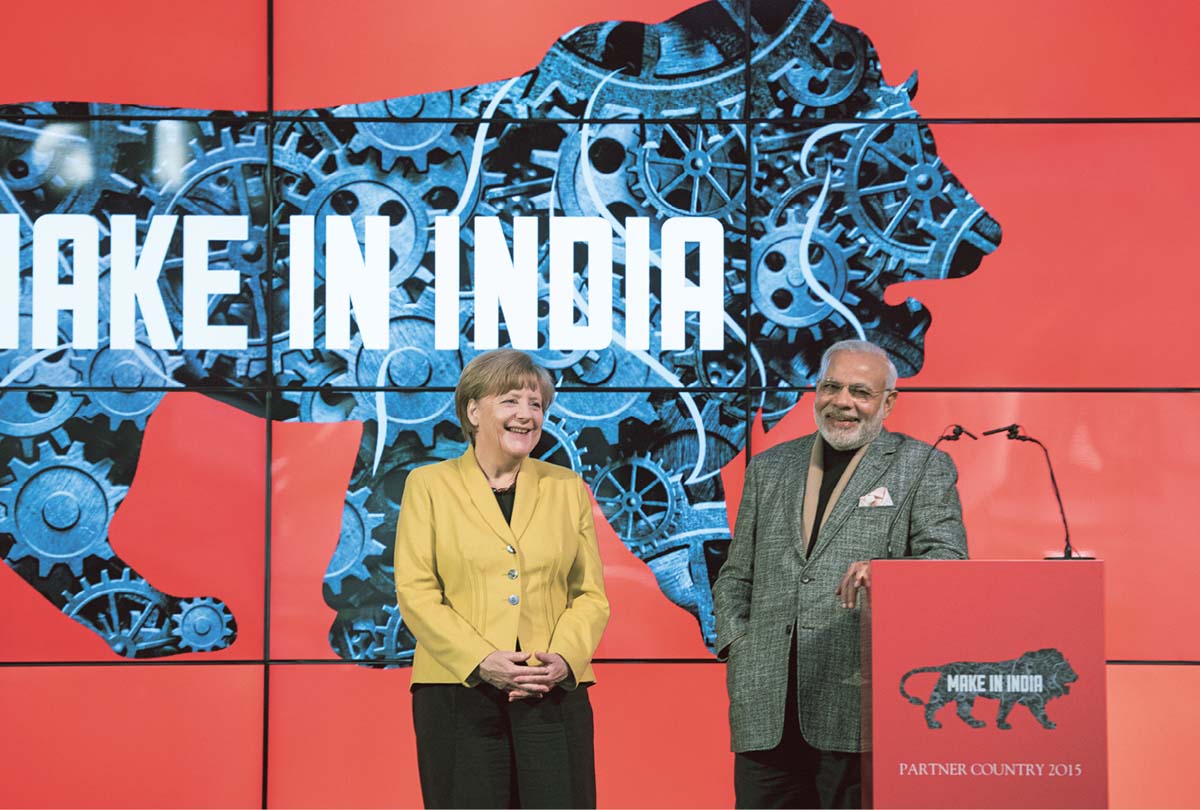 Narendra Modi steht an einem Rednerpult, neben ihm Angela Merkel. Im Hintergrund Scherenschnitt eines Löwen vor einer roten Wand, im Inneren des Löwen viele Zahnräden und der Schriftzug "Make in India".