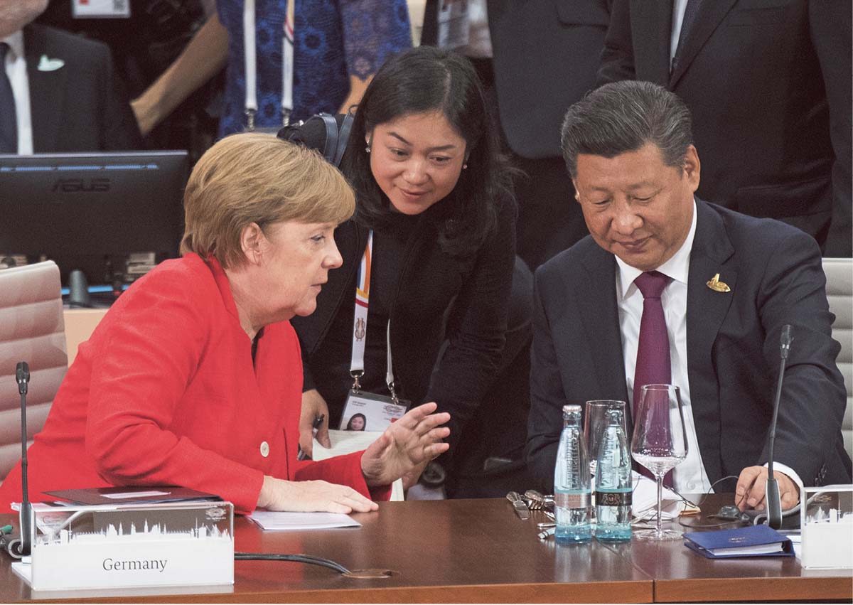 Angela Merkel sitzt neben Xi Jinping an einem Besprechungstisch, etwas ihnen steht eine asiatisch gelesene Frau, die sich zu ihnen hinunterbeugt.