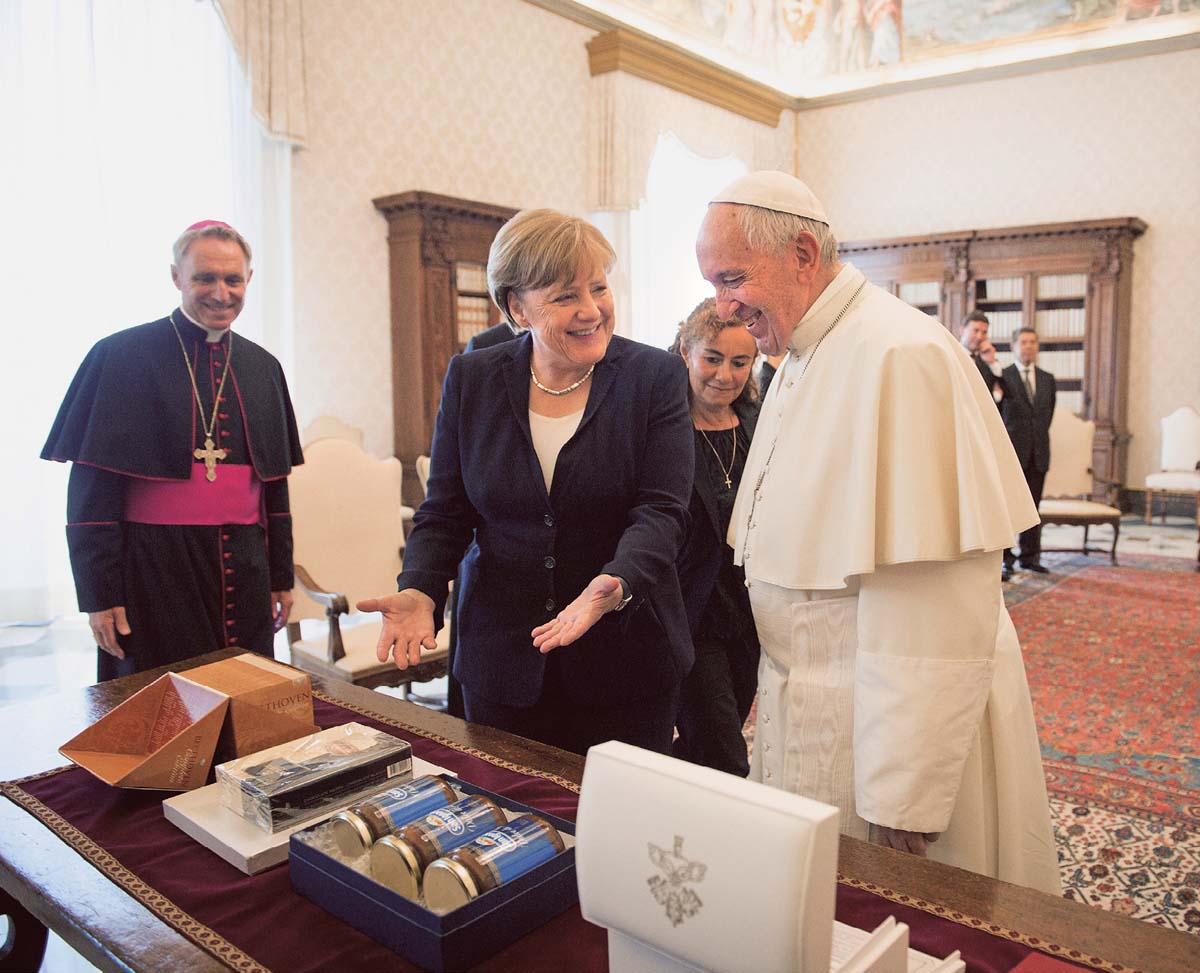 Angela Merkel steht neben Papst Franziskus und weist mit den Händen auf mitgebrachte Geschenke, die auf dem Tisch vor ihnen liegen. 