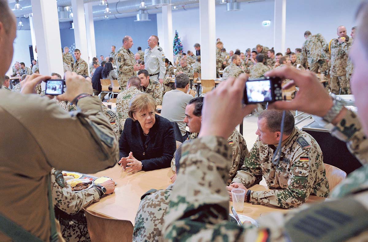 Angela Merkel in einem Raum voller Männer in Tarnkleidung. Sie sitzt am Tisch und spricht mit ihren Nachbarn und wird währenddessen von mehreren Männern fotografiert oder gefilmt.