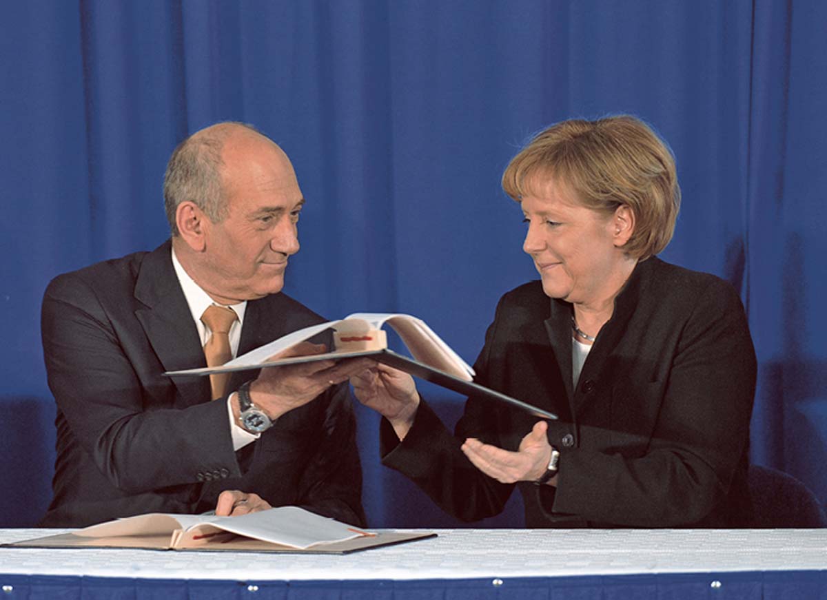 Ehud Olmert und Angela Merkel sitzen an einem Tisch vor einem blauen Vorhang. Er hält ihr einen aufgeschlagenen Aktenordner hin.