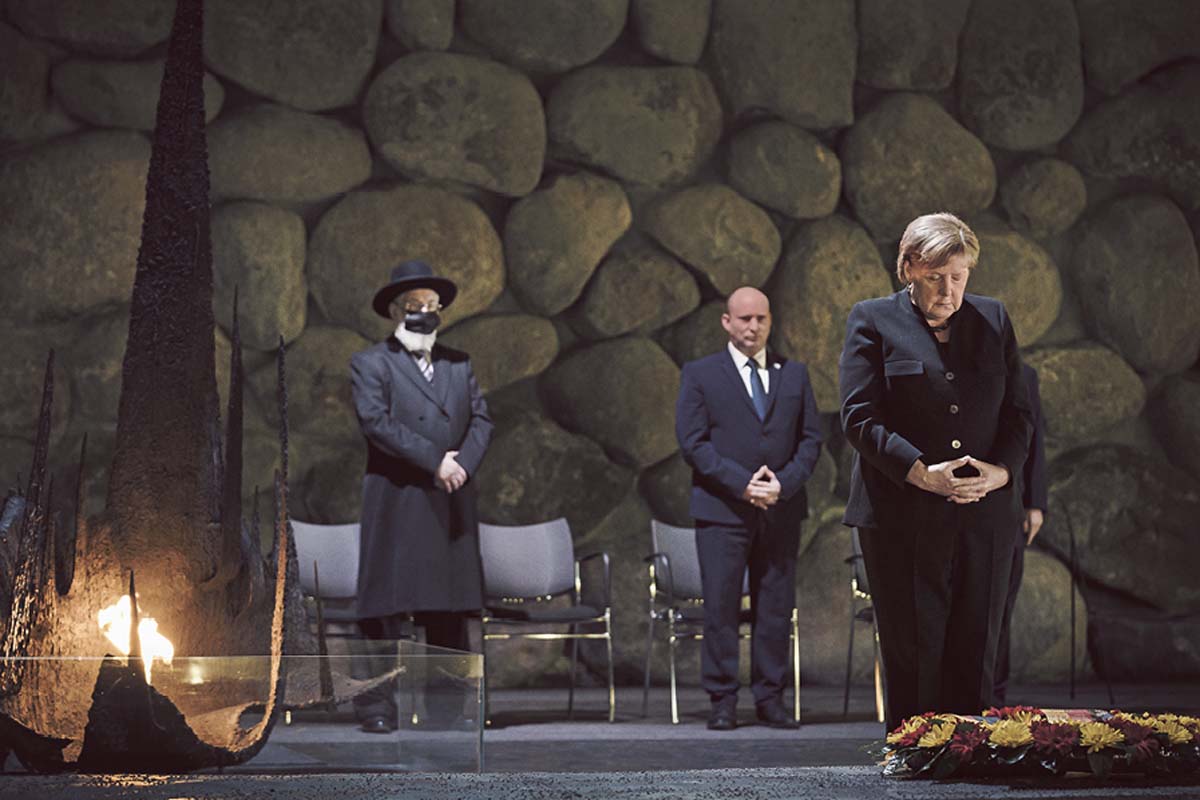 Angela Merkel steht in einem dunklen Raum, blickt zu Boden, hält die Hände zu einer Raute. Hinter ihr stehen Naftali Bennet und ein weiterer Mann vor mehrern Stühlen. Diese stehen vor einer Wand aus enormen Felsbrocken,