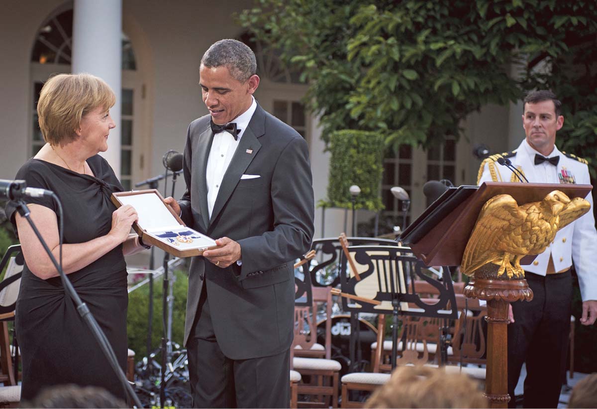 Angela Merkel in einem schwarzen ärmellosen Kleid steht neben Barack Obama im Smoking, der ihr eine Kiste hinhält, in der sich eine Medaille befindet. Vor ihr steht ein Mikrofon, neben ihm ein Rednerpult. Im Hintergrund ist das Weiße Haus zu erkennen. 