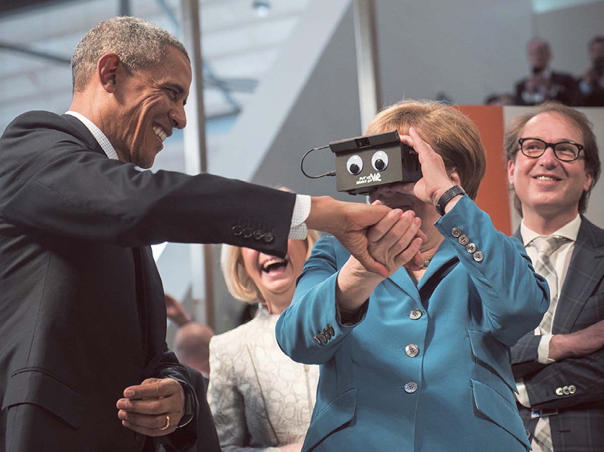 Angela Merkel hält die Hand des neben ihr stehenden Barack Obama. Mit der anderen Hand hält sie ein Gerät vor sich, in das sie hineinblickt. Im Hintergrund lachende Menschen.