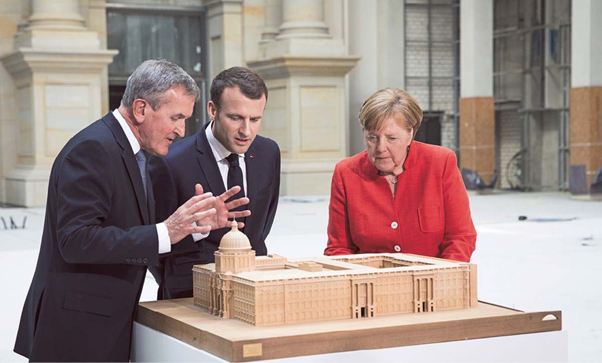 Angela Merkel steht neben Emmanuel Marcron, gemeinsam hören sie dem neben ihm stehenden Neil MacGregor zu. Sie blicken auf ein Miniaturmodell des Berliner Stadtschlosses. Im Hintergrund Säulen des Humboldt Forums