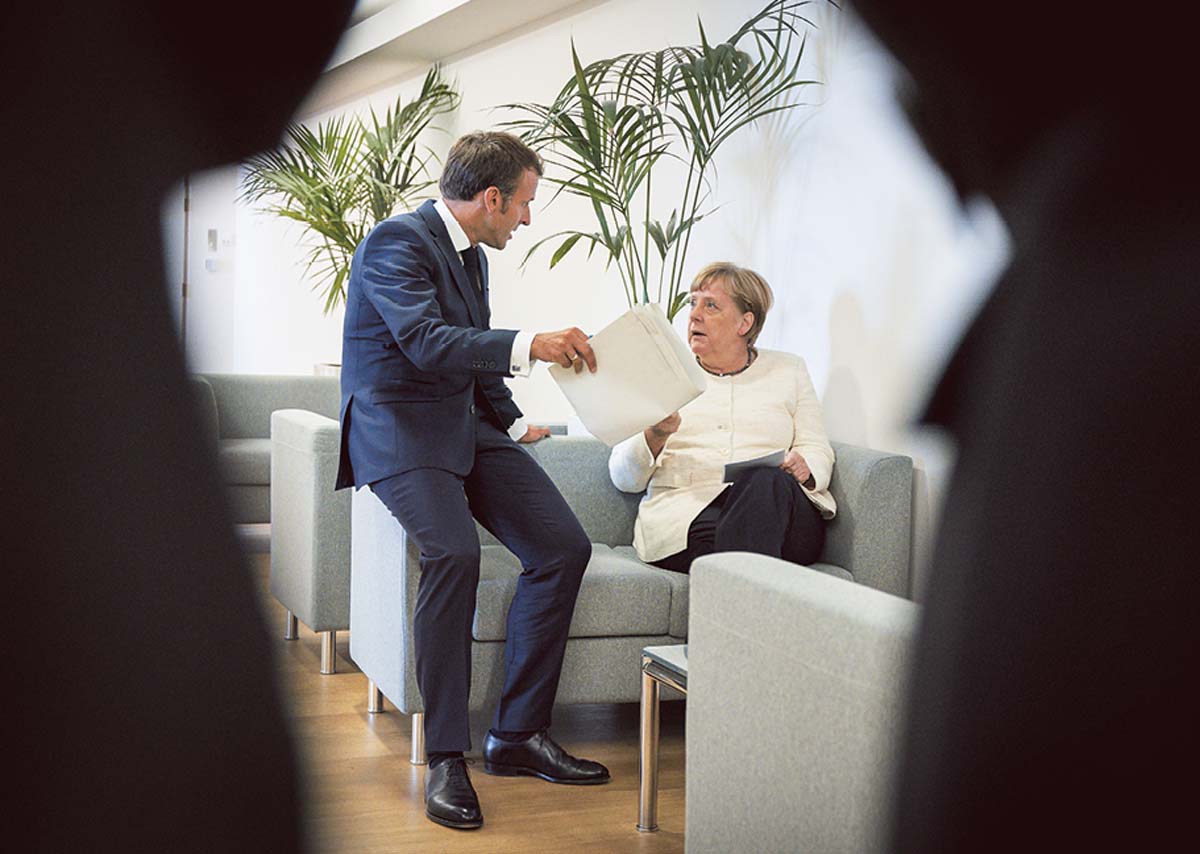 Blick durch ein Schlüsselloch auf Angela Merkel, die auf einem zweisitzigen Sofa sitzt. Emmanuel Macron lehnt an der Kante und hält ihr ein Papier hin.