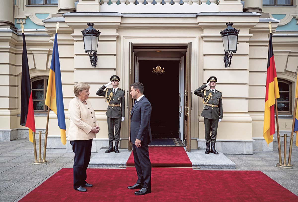 Wolodymyr Selenskyj und Angela Merkel stehen einander auf einem roten Teppich gegenüber. Im Hintergrund salutierende Soldaten vor einer imposanten Tür eines Palastes.