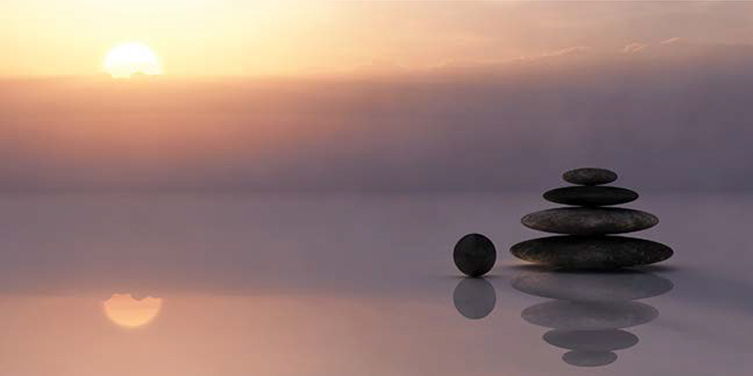 A stack of pebbles is shown with a sunny backdrop. The stack of pebbles is focused more compared to the sun.