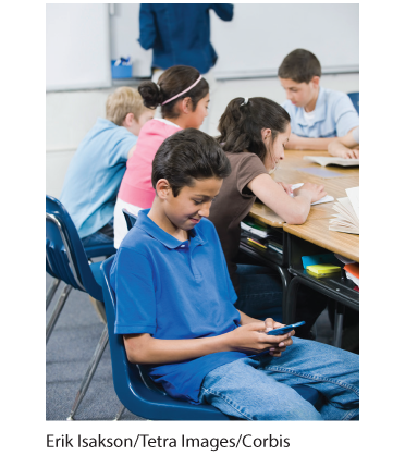 This is an image of a boy reading his smartphone with several other boys and girls sitting around a table reading books.