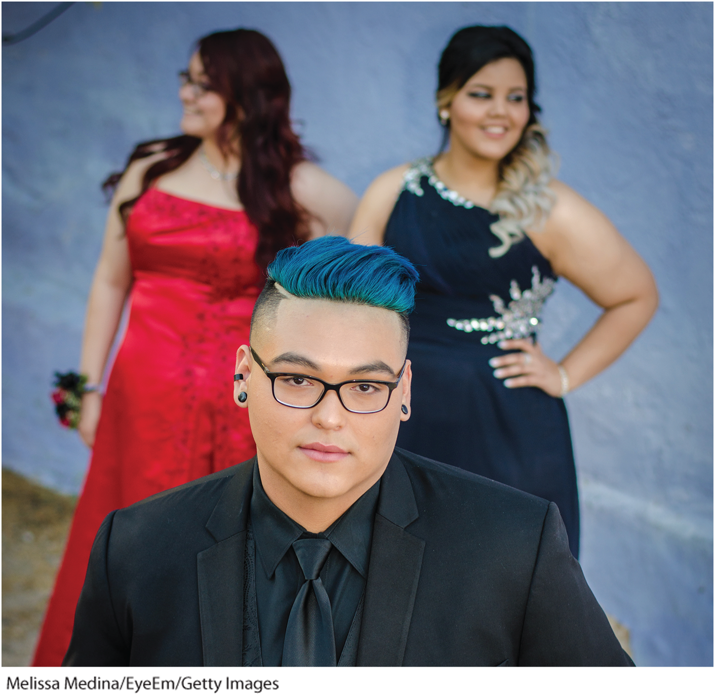 A photo shows a bespectacled young man with dyed hair in the foreground and two young women in the background.