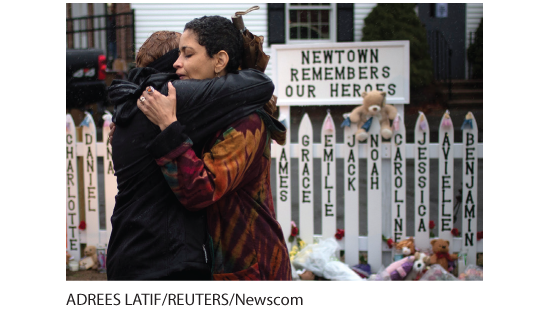 A photo shows a woman hugging another woman. At the background a sign reads, Newtown remembers our heroes. A picket fence has the names of the victims from the Newtown mass shooting.