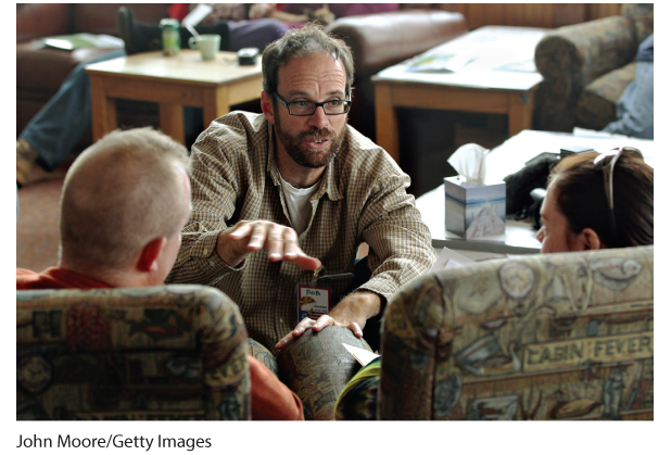 A photo of a man speaking to a man and a woman who are sitting across him.