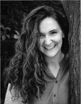 A woman with long, dark curly hair is smiling at the camera and leaning against a tree