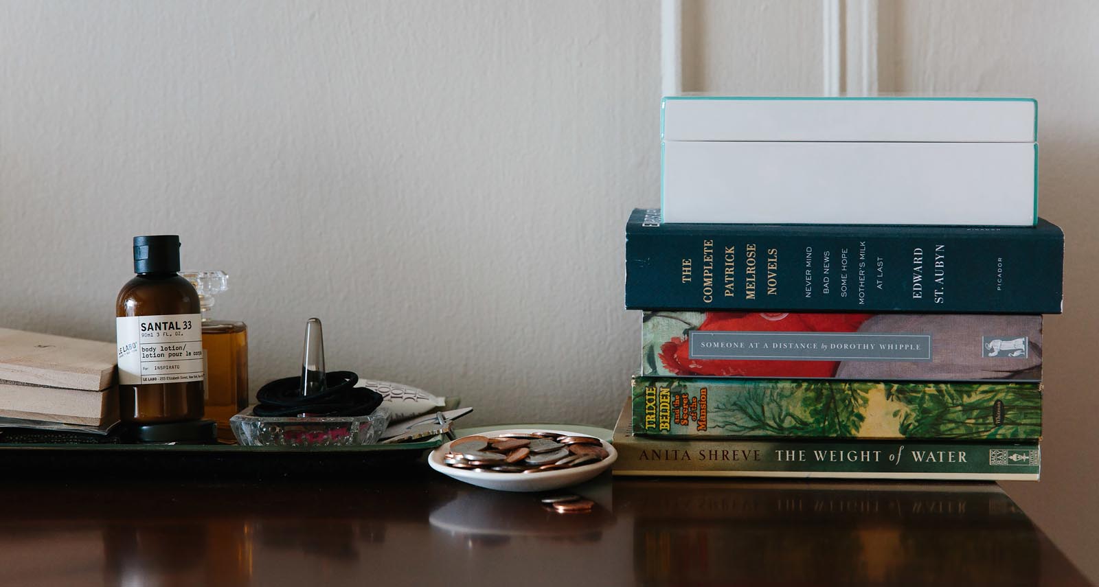 Photo of a stack of books shares a tabletop with a dish of coins, perfume, hair-ties, and other daily items.