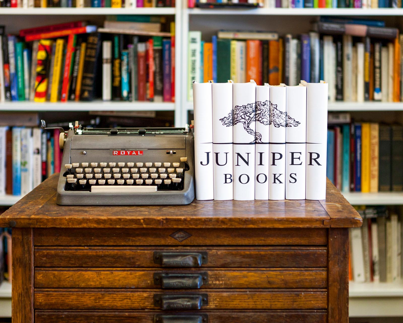 Photo of a set of shallow drawers in front of a bookshelf with a typewriter and a set of books jacketed with the Juniper Books logo.