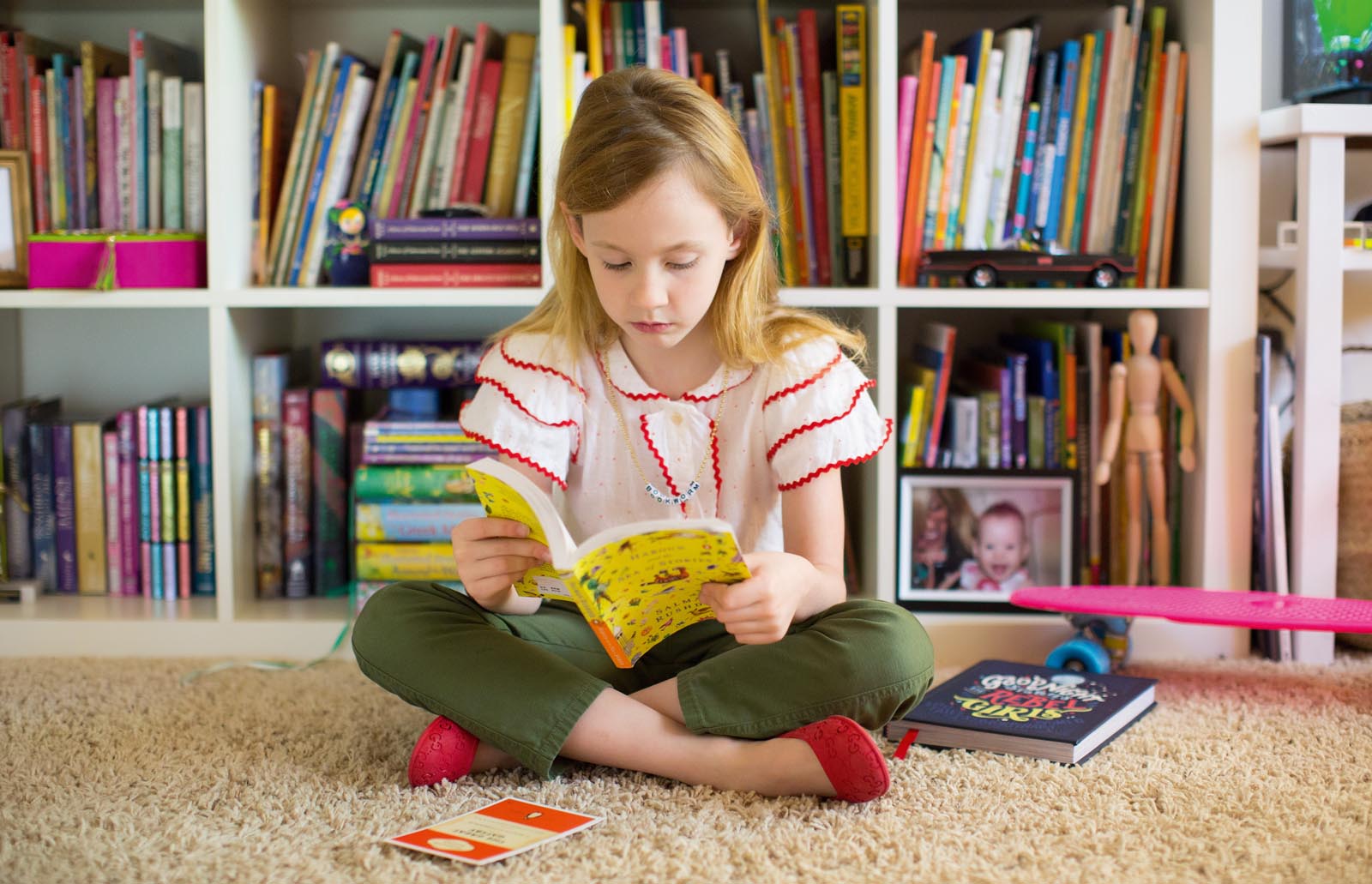 Photo of a girl reading a paperback in front of a set of book-laden shelves.