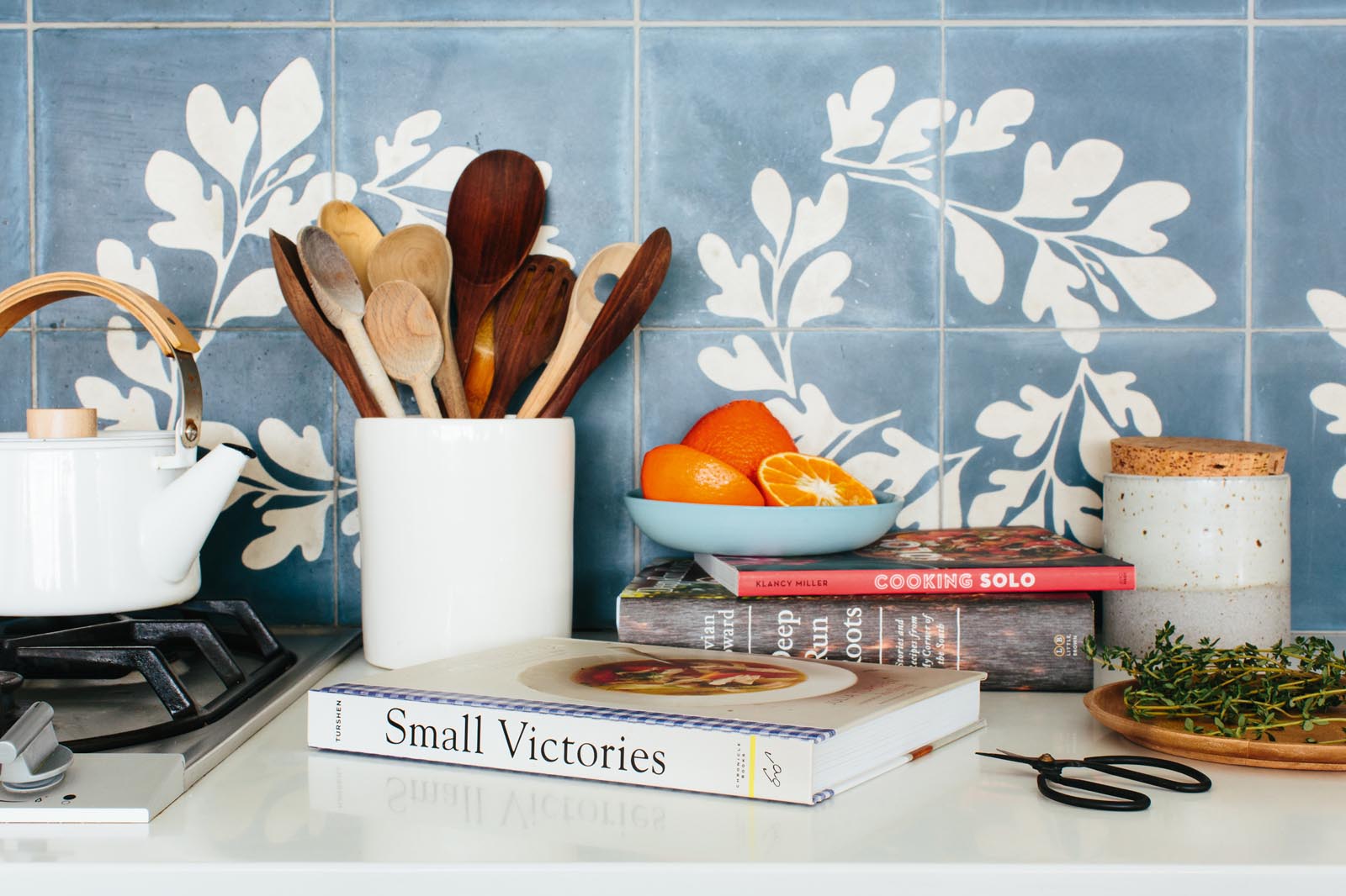 Photo of a kitchen counter featuring cookbooks, herbs, utensils, and a teapot.