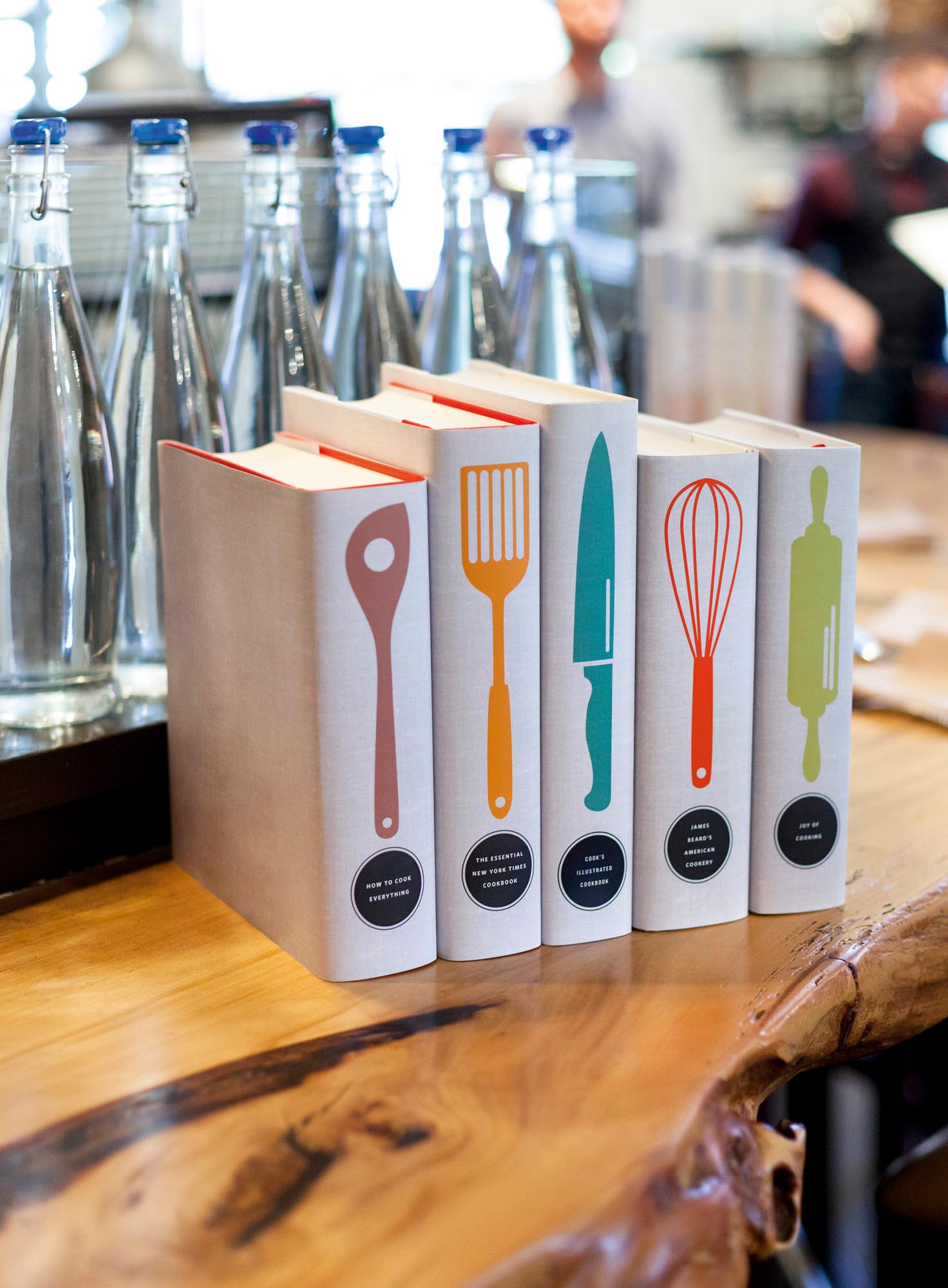 Photo of a collection of books jacketed with colorful kitchen utensils on a natural wood counter.