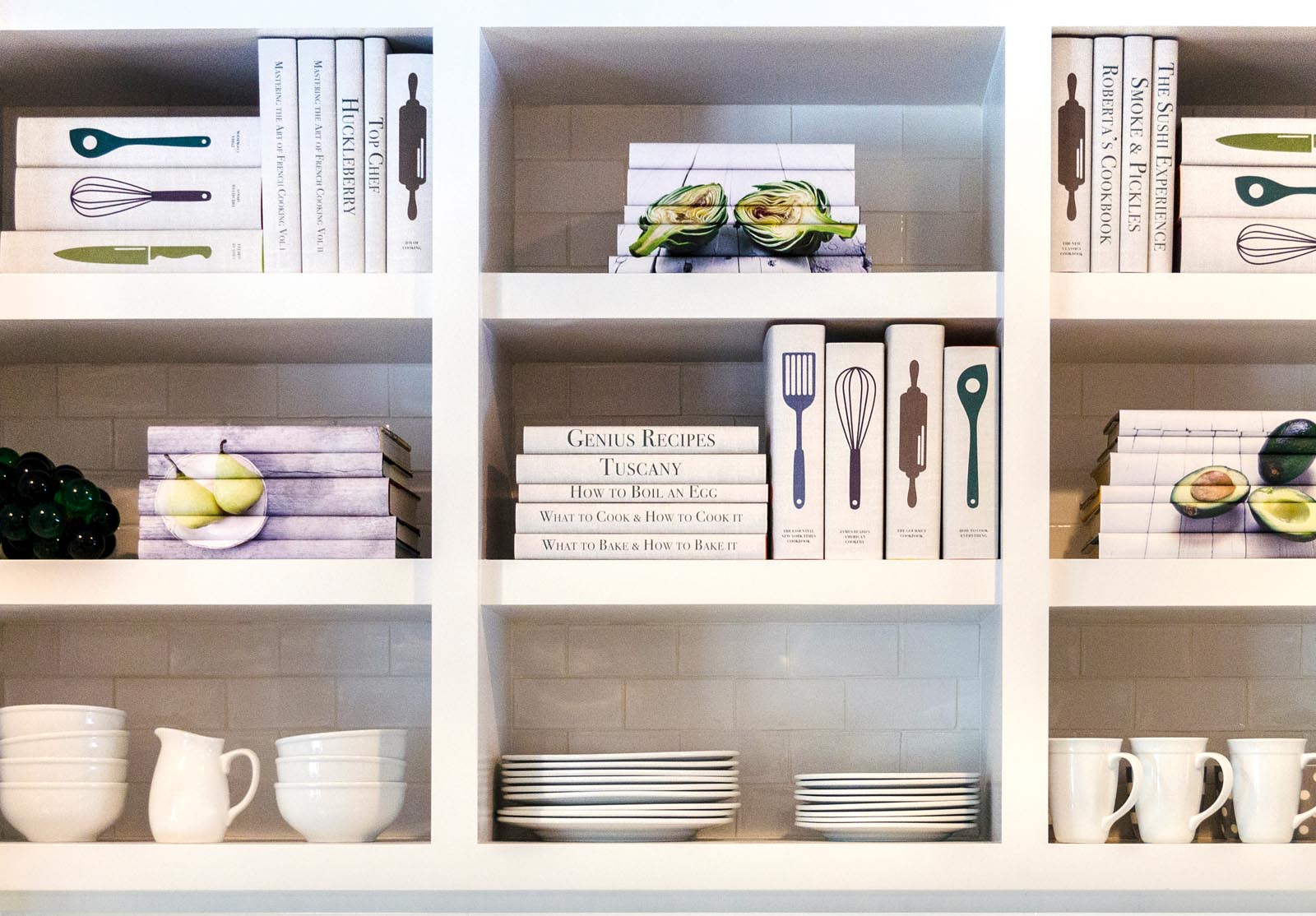 Photo of collections of custom-jacketed books in light colors on white shelves along with white dishes.