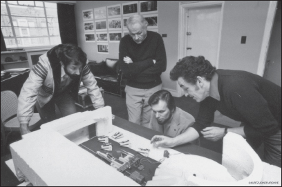 Cinematographer Peter Suschintzky, artist Ralph McQuarrie, Gary Kurtz, and production designer Norman Reynolds discuss an early scene in The Empire Strikes Back over a maquette of the Rebel Base hangar on Hoth.