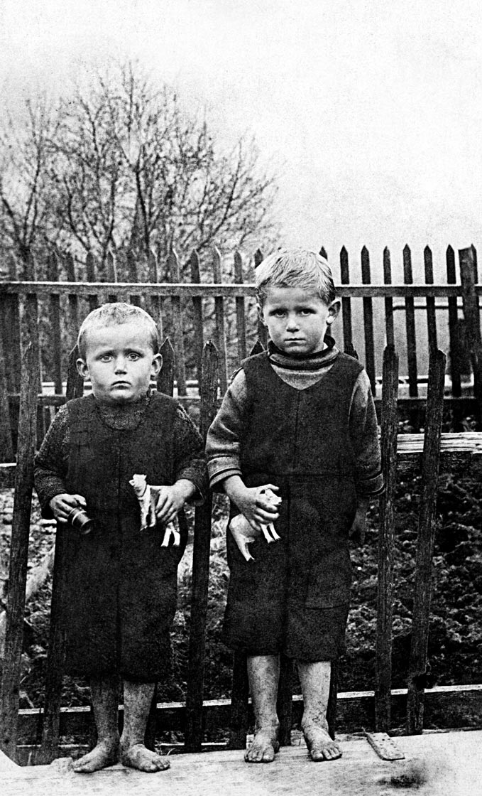 Zwei Jungen stehen vor einem Zaun, sie sind barfuß, haben schmutzige Füße und schauen sehr ernst in die Kamera. Jeder hat eine kleine holzgeschnitzte Kuh in der Hand.