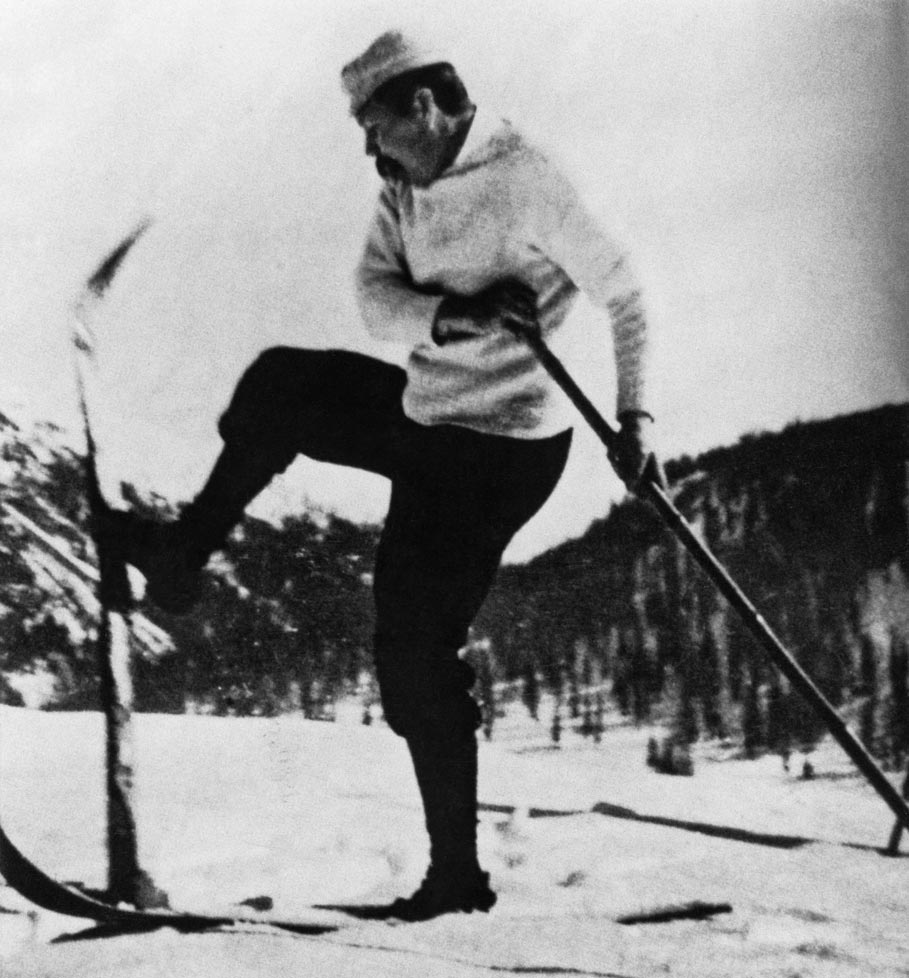 Eine Großaufnahme von Sir Arthur Conan Doyle im hellen Pullover mit Mütze. Er versucht seinen langen Ski zu wenden, indem er sich mit einem Stock abstützt.