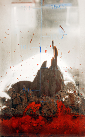 Close-up of ice cream, chocolate cake scraps, and red food coloring layered against a clear plastic wall