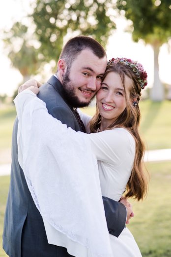 Image is of the author, Brianna Campbell, and her husband, Scott Campbell, on their wedding day. Brianna is wearing a white dress with long wrapping sleeves and has her brown hair down and wavy. She is wearing a pink and blue flower crown and hugging her husband, who is wearing a grey suit. He has a short beard and mustache and buzzed hair. They are standing outside in a green field with trees in the background.