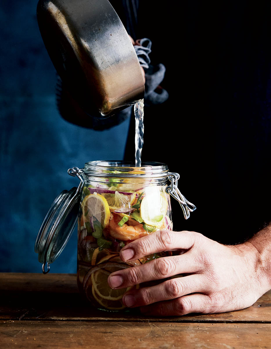 A chef pouring freshly boiled brine into a mason jar full of herbs and shrimp for pickling.