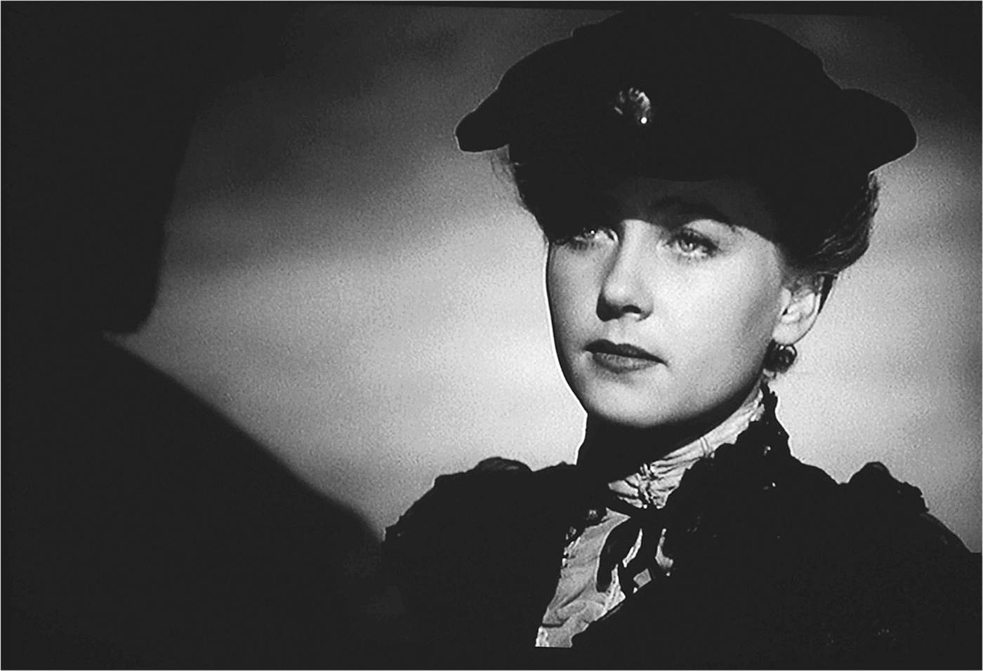 Head shot of a woman, with a dark background. She wears a hat and does not seem to be focusing on anything.