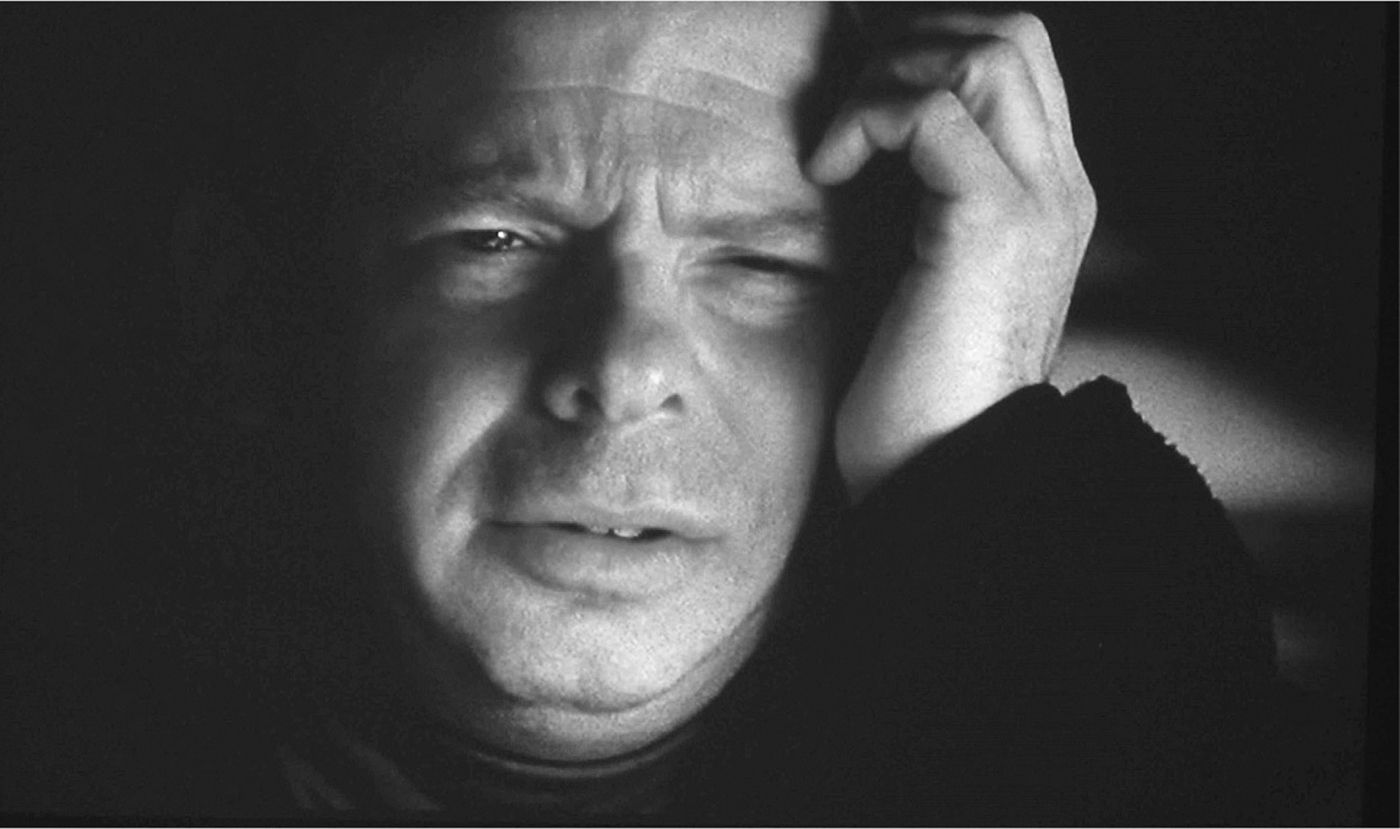 Head shot of a man, with a dark background, his face pained, his cupped hand at his temple. 