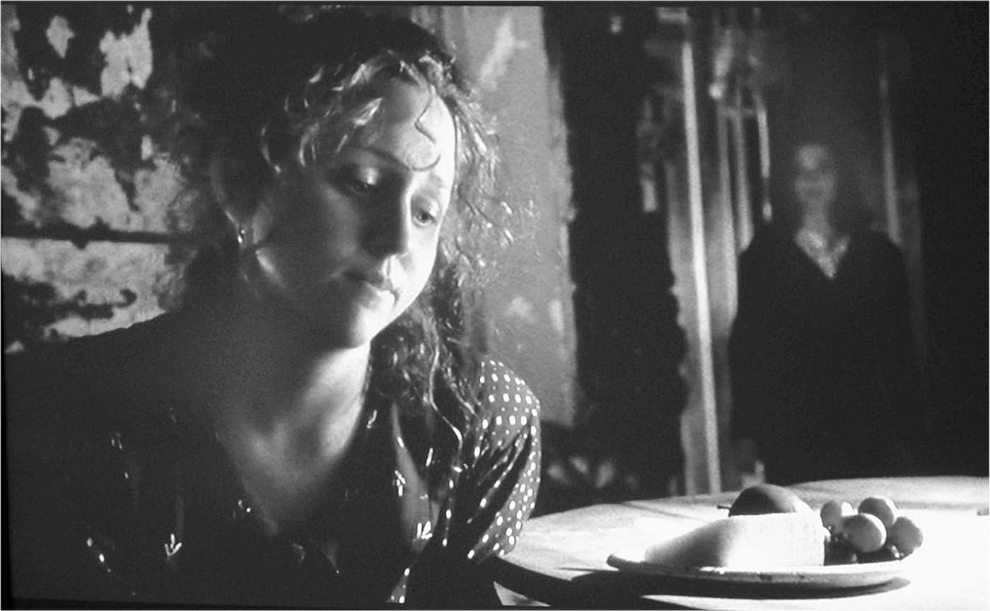 Head shot of a woman looking down at a plate of fruit and cheese on a table. In the dark background, another woman looks at her.