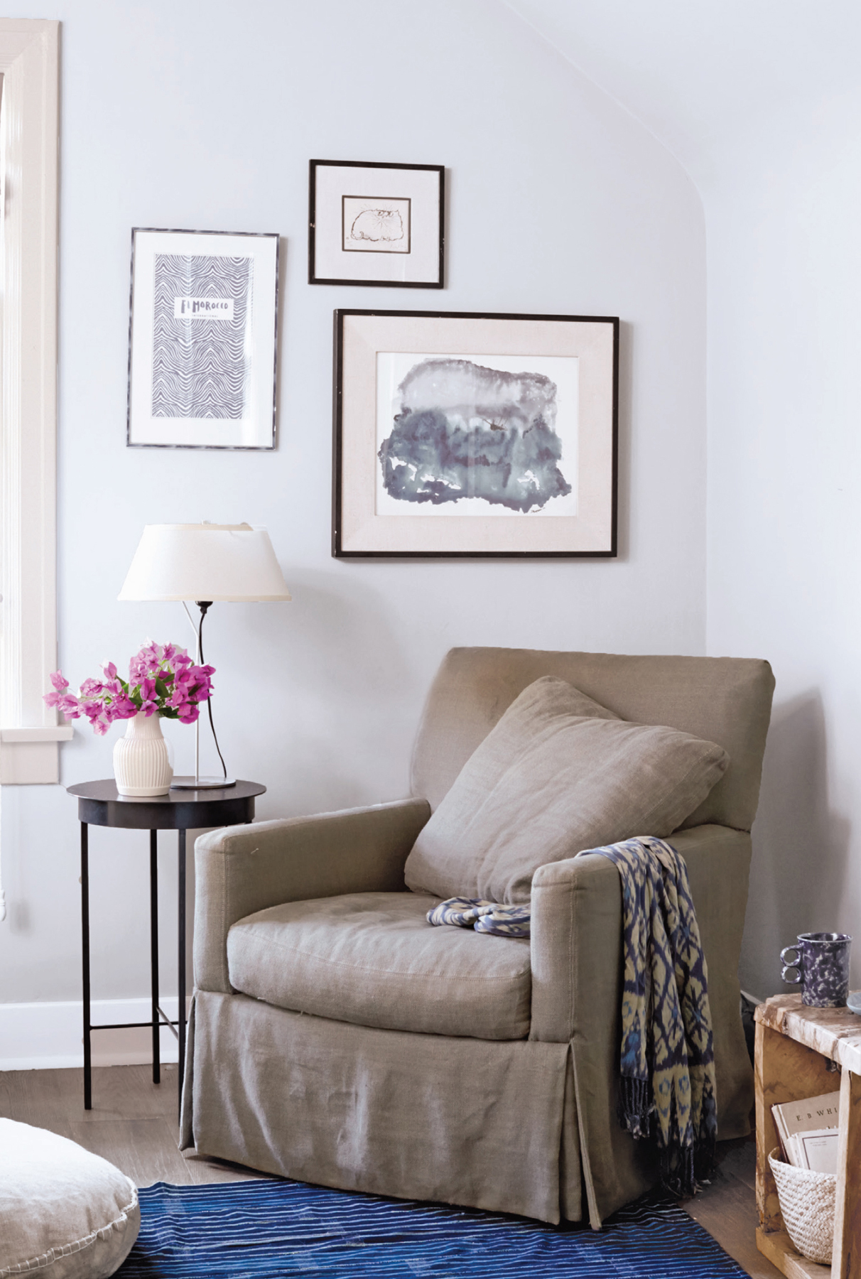 A chair, side table, and art create a quiet reading nook with a throw waiting for you to snuggle in. The pink flowers intersect the vertical line of the lamp, while the basket at right draws your eye quietly, adding texture and depth.