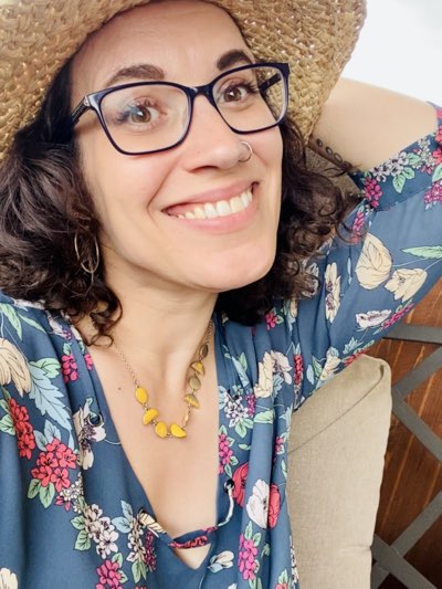 Poppy Parkes author photo: Curly-haired brunette with glasses wearing hat and smiling