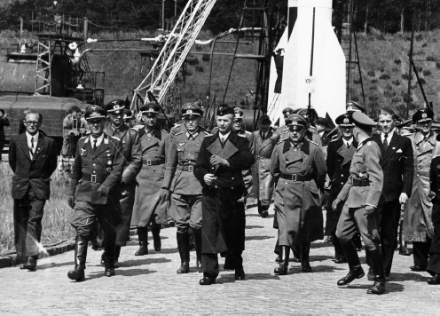 With an A-4 (V-2) missile in the background, von Braun (at right in dark suit) accompanies a contingent of top ...