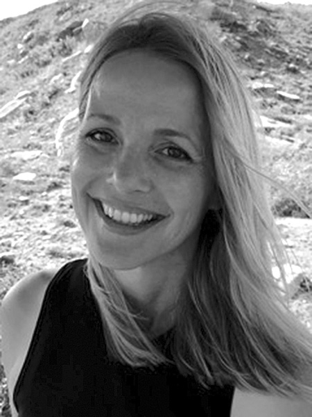 A black-and-white photograph of Barbara Radecki with long hair and a black sleeveless top on with a background of a rocky, grassy hill behind her. She has a wide smile and joyful eyes.