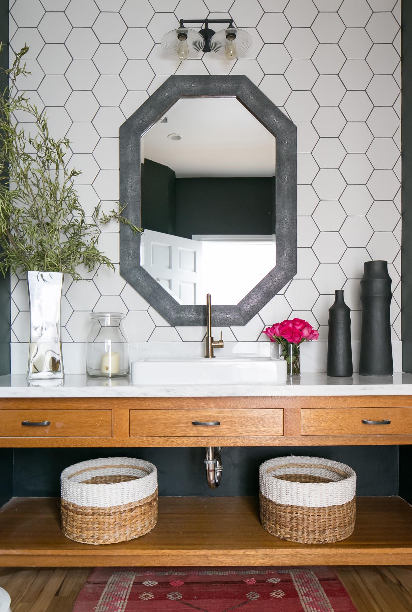 Hexagonal bath tiles framed by an eight-sided parlor mirror backstop a composition vanity of natural wood and marble.